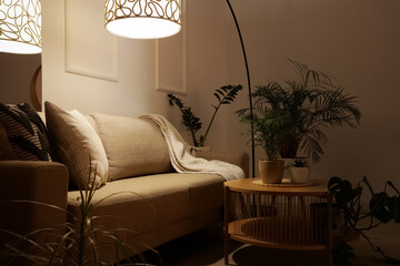 Interior of stylish living room with cozy grey sofa, coffee table, houseplants and glowing lamps at...