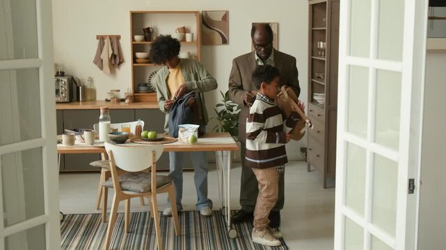 Dolly in shot of senior African American man sending young grandsons to school giving kids bags with apples and sandwiches for lunch, family morning routine in kitchen