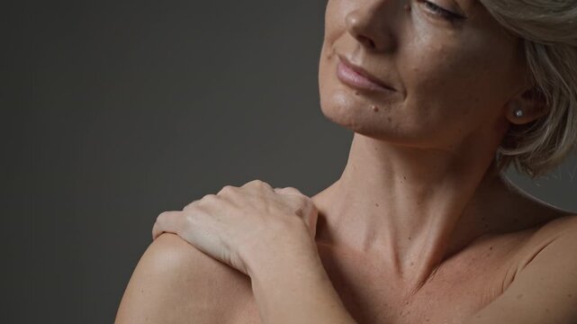 Close up shot of middle-aged Caucasian blonde woman posing for camera with bare shoulders on minimal grey background, natural beauty concept