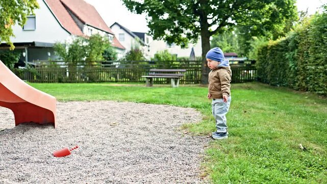 A 2 year old boy in a jacket plays outside on a warm autumn day. Toddler plays on a playground in a city park. The child breathes fresh air and explores the world around him.