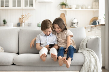Two diverse little children using phone sitting on cozy couch