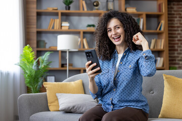 Obraz premium Young woman with curly hair sitting on a couch, enthusiastically cheering while looking at her smartphone and celebrating a victory, success, or positive news from an app or online game