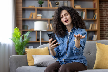 Obraz premium Woman with curly hair sitting on sofa, looking perplexed and making a gesturing hand, clearly expressing frustration and confusion while holding and looking at her smartphone