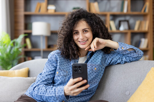 Smiling young woman with curly hair relaxing on a sofa, holding a smartphone and looking at the camera, enjoying connectivity and casual leisure time at home