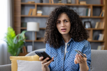 Young woman feeling stressed and confused, holding a smartphone and credit card, facing an online shopping payment error or financial problem in a home setting