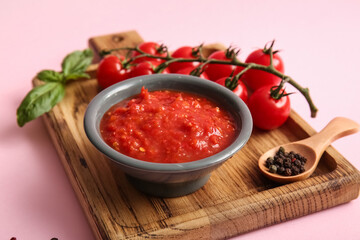 Board with bowl of tasty tomato sauce and ingredients on pink background, closeup