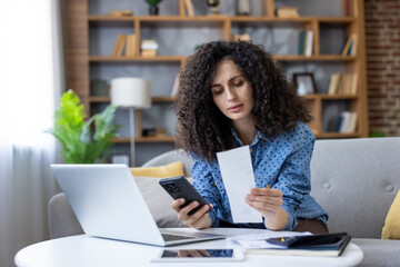 Woman holding a receipt and smartphone, comparing expenses while managing her budget at home,...