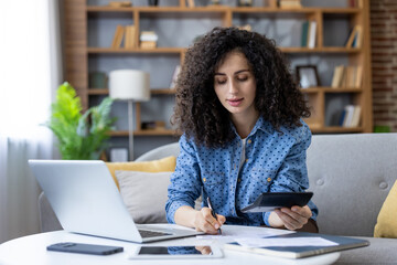 Young woman managing personal finances and household budget, calculating expenses and reviewing financial paperwork while working remotely from her comfortable home office