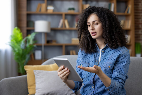 Young woman with curly hair sitting on a sofa, gesturing and speaking into a tablet during a remote video conference for work, education or online collaboration