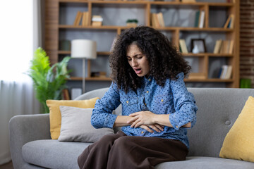 Fototapeta premium Woman sitting on a grey couch at home, experiencing severe abdominal pain or period cramps, holding her stomach with both hands and expressing discomfort