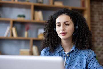 Woman with curly hair wearing a headset looking at a laptop screen, engaging in remote work,...