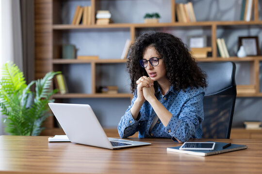 Worried woman with curly hair feeling overwhelmed and contemplating while working on her laptop at a wooden desk in a home office environment, dealing with stress and remote work challenges