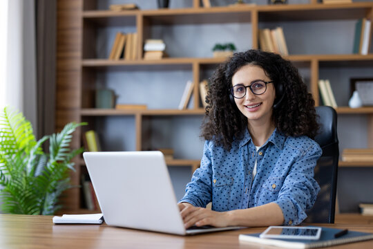 Young woman with curly hair and glasses smiling at camera while working from her home office, typing on a laptop with headset during a video call or virtual meeting, confident and cheerful