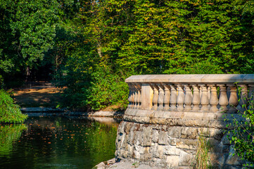 Fototapeta premium Stone Observation Deck Reflected In Calm Park Pond During Sunny Late Summer Afternoon, Nature Background