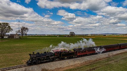 A historic steam train moves along its track, releasing clouds of steam. The surrounding fields are lush and green, while fluffy clouds float in the blue sky.