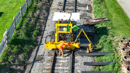 Two workers focus on maintaining train tracks in a rural area. The yellow machine helps them lift and position wooden planks as they ensure safety and functionality.