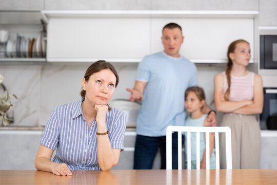 Family quarrel. Sad mom is sitting near table in kitchen and listens to abuse of angry husband. Children took fathers side and hugged dad during difficult unpleasant conversation between parents