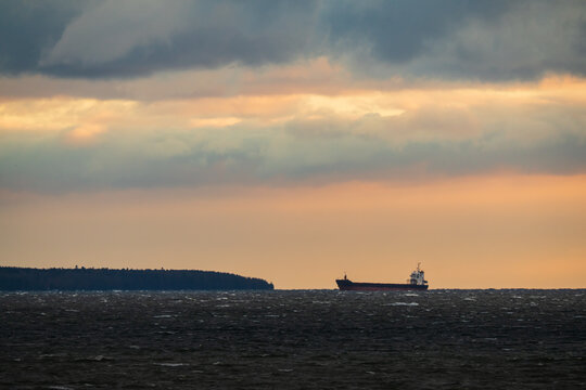 Cargo ship sailing across the rough waters of the Baltic Sea near the Estonian coast under a dramatic, warm-lit sunset sky and dark clouds.