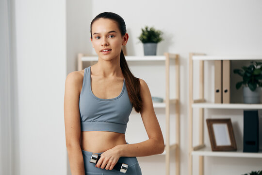 Woman in athletic wear holding a dumbbell during a home workout, bright room with shelves, plants and framed art, showcasing personal training, fitness routine, and concentration. - Powered by Adobe