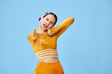 Energetic woman in orange fitness outfit dancing and stretching, wearing headphones against a bright blue studio background. Dynamic movement, cardio workout vibe, sporty style, vibrant energy and