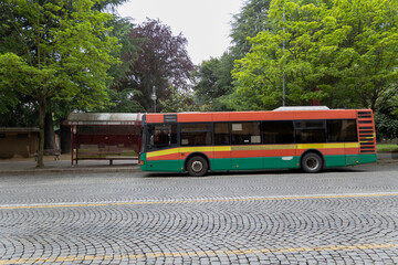Urban bus with a colorful livery stopped at the bus stop, surrounded by trees on a summer day. 