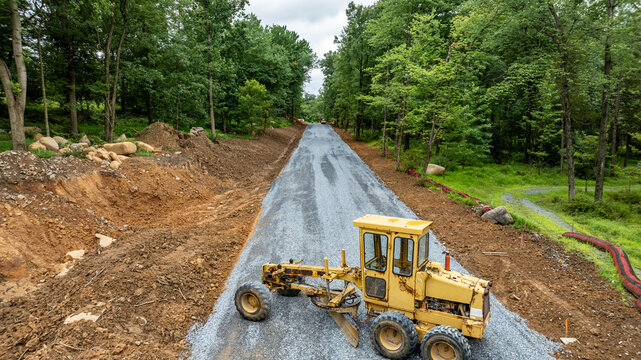 Heavy machinery is grading a recently constructed gravel road surrounded by trees and dirt piles. The work site shows preparation for road development in a natural setting.