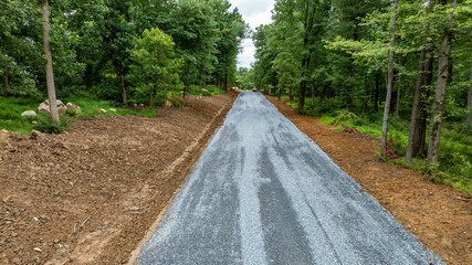 Construction is underway for a new gravel road winding through a lush green forest. Trees line the path, and earth has been cleared on both sides to improve accessibility and safety.