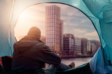 Homeless man sitting in a blue light tent by the entrance and looking across river on a block of...