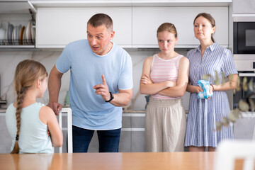 Obraz premium Family scene in kitchen, unpleasant conversation. Girl with hair in braids and crossed arms on chest stands in front of parents and listens to instructions about her bad behavior