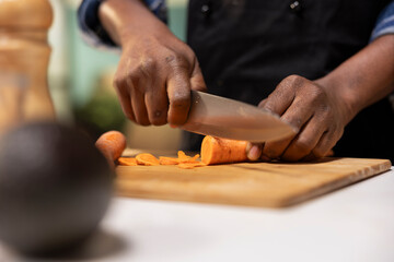 Close up of african american man cutting carrots for a fresh salad, preparing organic ingredients on wooden board at the kitchen counter. Person chopping colorful vegetables for a healthy meal.
