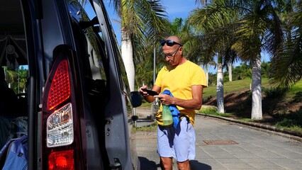Mature man cleaning his car mirror with a cloth and spray bottle, holding a smartphone, actively engaged in vehicle maintenance parked outdoors on a sunny day