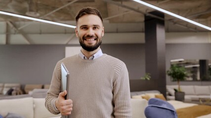 Young man standing in a contemporary showroom. He is holding a laptop and smiling at the camera, showing confidence