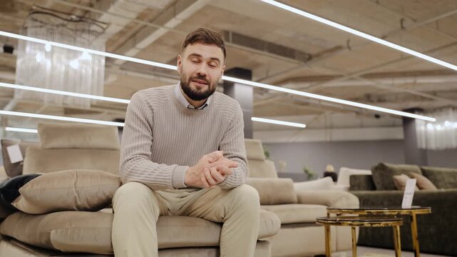 Young man talking to the camera, sitting on a modern sofa set, recording video in a contemporary furniture showroom