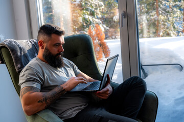 Freelancer with tattoos and a beard working on a laptop in an armchair by a window, enjoying the...