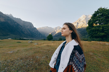 Fototapeta premium Woman portrait in mountains meadow, nature outdoors and travel landscape setting with thoughtful expression, cozy patterned coat and wide valley view at golden hour.