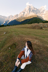 Fototapeta premium Woman in autumn sweater sits in a mountain meadow, nature landscape with high peaks and soft light, travel and hiking scene, peaceful outdoor solitude and scenic horizon.