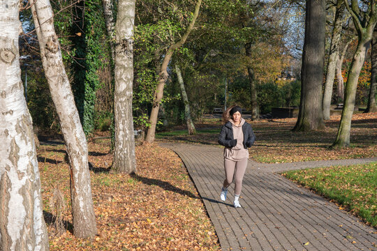 Young woman jogging alone on a paved park path, engaging in an outdoor activity during autumn, staying fit and promoting wellness with fallen leaves on the ground