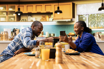 Joyous boyfriend and girlfriend sharing breakfast pastries and coffee at home. Laughing and bonding over casual conversation, watching funny videos on smartphone for a candid cute moment.