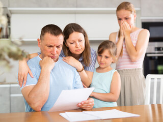 Fototapeta premium Caring wife and two teenage daughters calming upset puzzled father sitting with papers at table in home kitchen