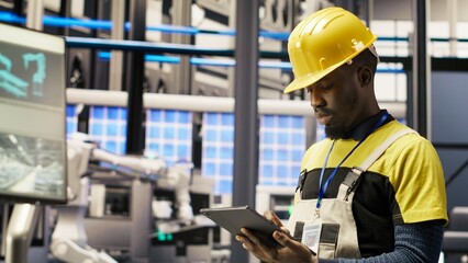 Smart factory mechanic using tablet to analyze IoT sensors data before modifying gear. African american man in industry 4.0 plant performing system diagnostics to reduce downtime, camera B