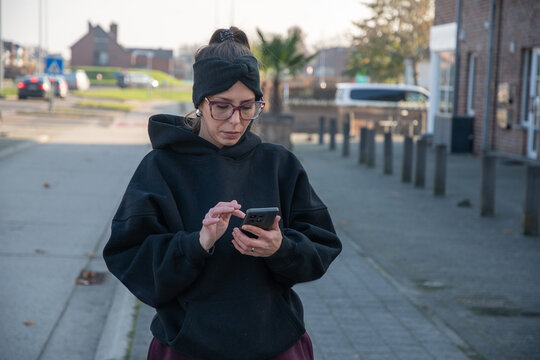 Young woman on an urban sidewalk intently using her smartphonetexting, browsing apps and social media, connected and focused on digital communication and daily mobile routines - Powered by Adobe