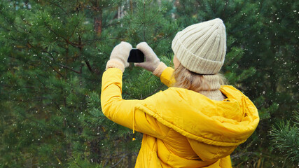 Young woman taking picture on phone Christmas tree in winter park, girl with smartphone in forest
