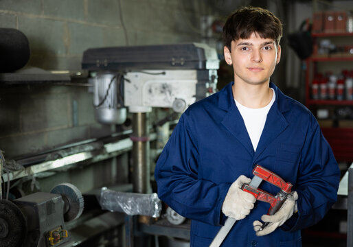 Young guy, professional technician with clamp in hands, wearing blue work coat and gloves, posing in industrial metal workshop, looking confidently and friendly at camera..