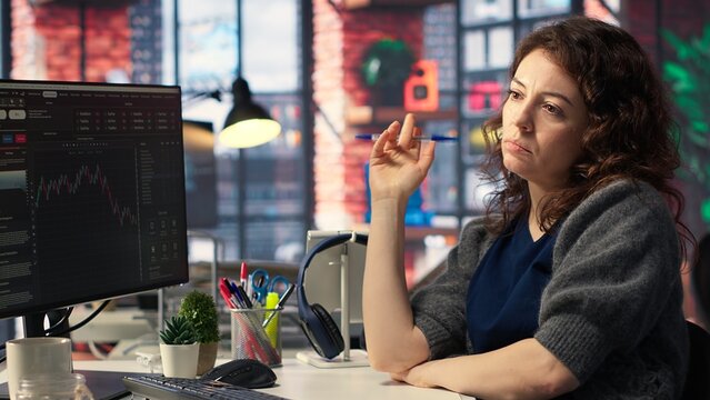 Focused woman studies stock trading charts at computer setup, browsing through financial analysis tools. The trading platform provides statistics, forex trends and index for profitable investments