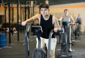 Dedicated active strong young man doing cardio workout on stationary bicycle as hard cross-training in gym indoors