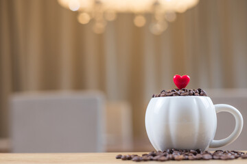 White cup filled with coffee beans and small red heart on wooden table with scattered beans and copy space on left