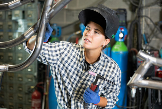 Enthusiastic young female welder in casual plaid shirt, protective gloves and helmet welding metal pipe structure in workshop, holding torch and carefully inspecting quality of weld seams