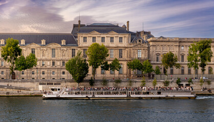 A tourist boat on waters of Seine River in Paris, France, in front of the Louvre Museum building with tourists enjoying the cruise along Paris's most beautiful historical axis