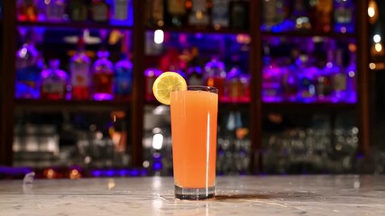 Orange cocktail with lemon slice in colorful bar. Tall glass of orange cocktail with lemon slice served on marble counter against neon bar background. - Powered by Adobe