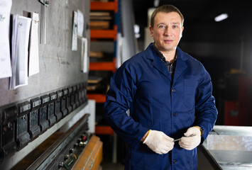 Experienced middle-aged male worker in blue workwear posing in metalworking workshop, surrounded by tools and machines, showcasing confidence and expertise in professional setting